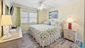 Ein Schlafzimmer mit Bett, Nachttisch, Schreibtisch, Deckenventilator, Fenster mit Vorhängen und einem Bild an der Wand.