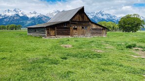 Un granero de madera rústico con techo inclinado, rodeado por un campo verde exuberante y montañas nevadas al fondo.