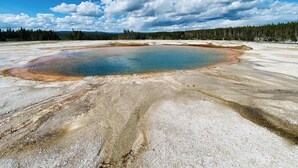 Una gran piscina geotérmica circular rodeada por un paisaje árido con poca vegetación y un bosque al fondo.