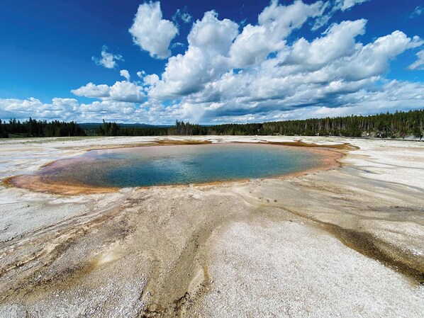 Una gran piscina geotérmica circular rodeada por un paisaje árido con poca vegetación y un bosque al fondo.