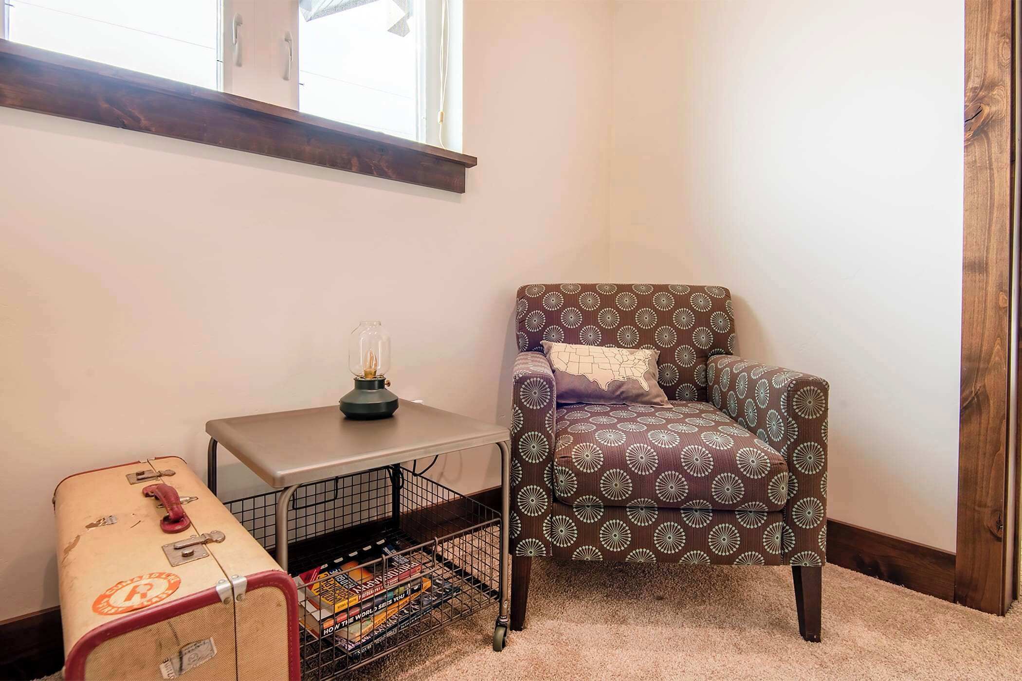 Een hoekzaak met een gestreept fauteuil, een klein tafeltje met een lamp, een koffer en een draadrek met boeken.