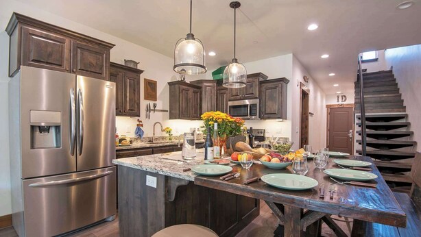Una cocina con una isla grande, electrodomésticos de acero inoxidable, gabinetes de madera e iluminación colgante, y una escalera.