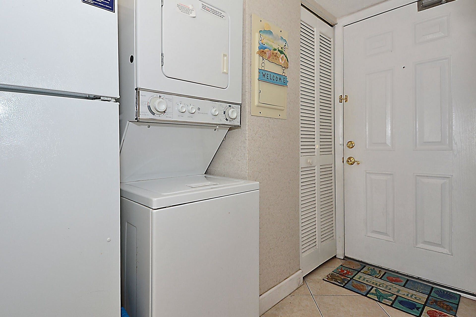 A laundry area with a washing machine and dryer, a welcome sign, and a door with a beach-themed mat.