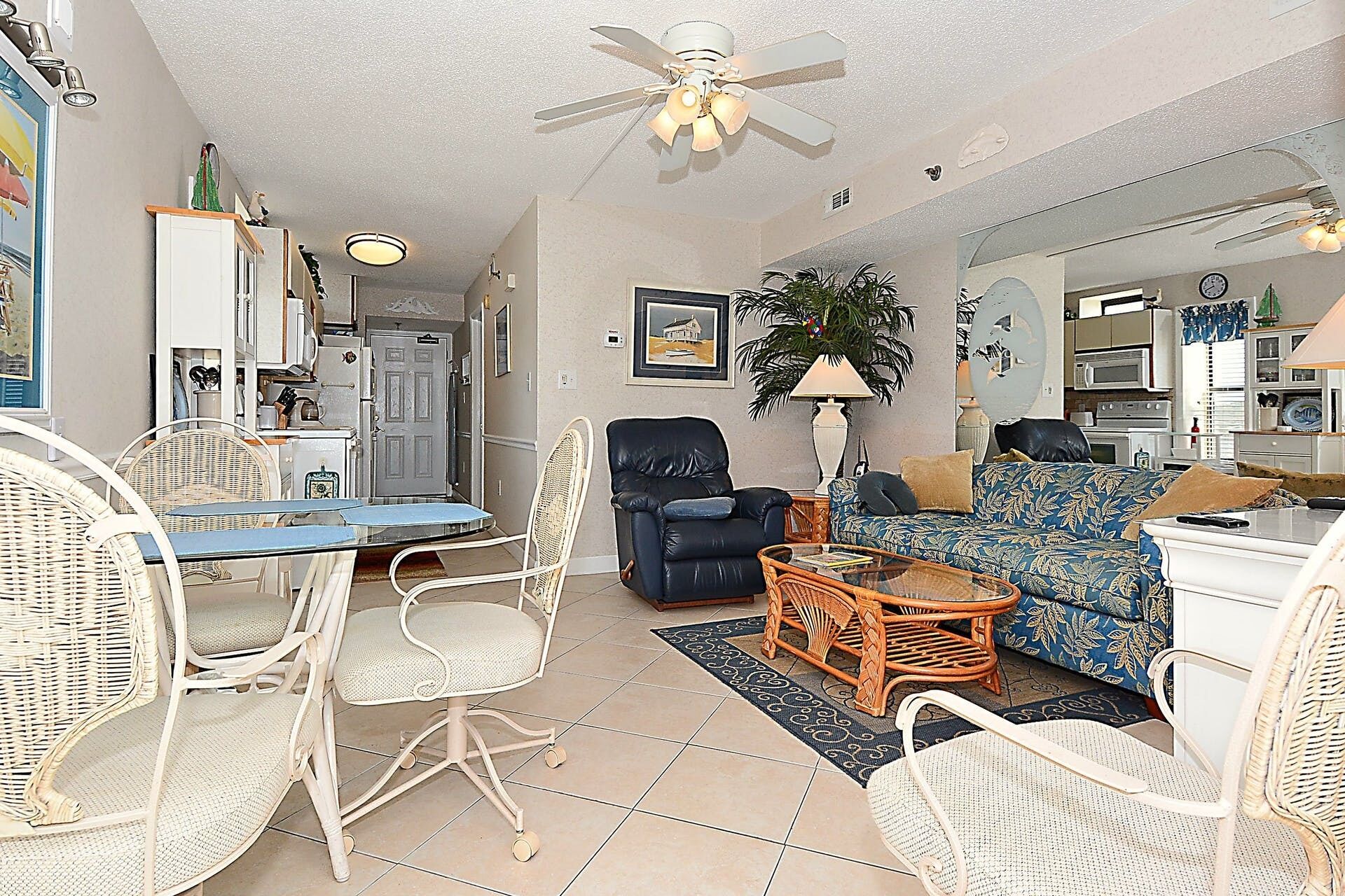 A living room with wicker furniture, a ceiling fan, a dining area with a table and chairs, and a kitchen area with appliances.