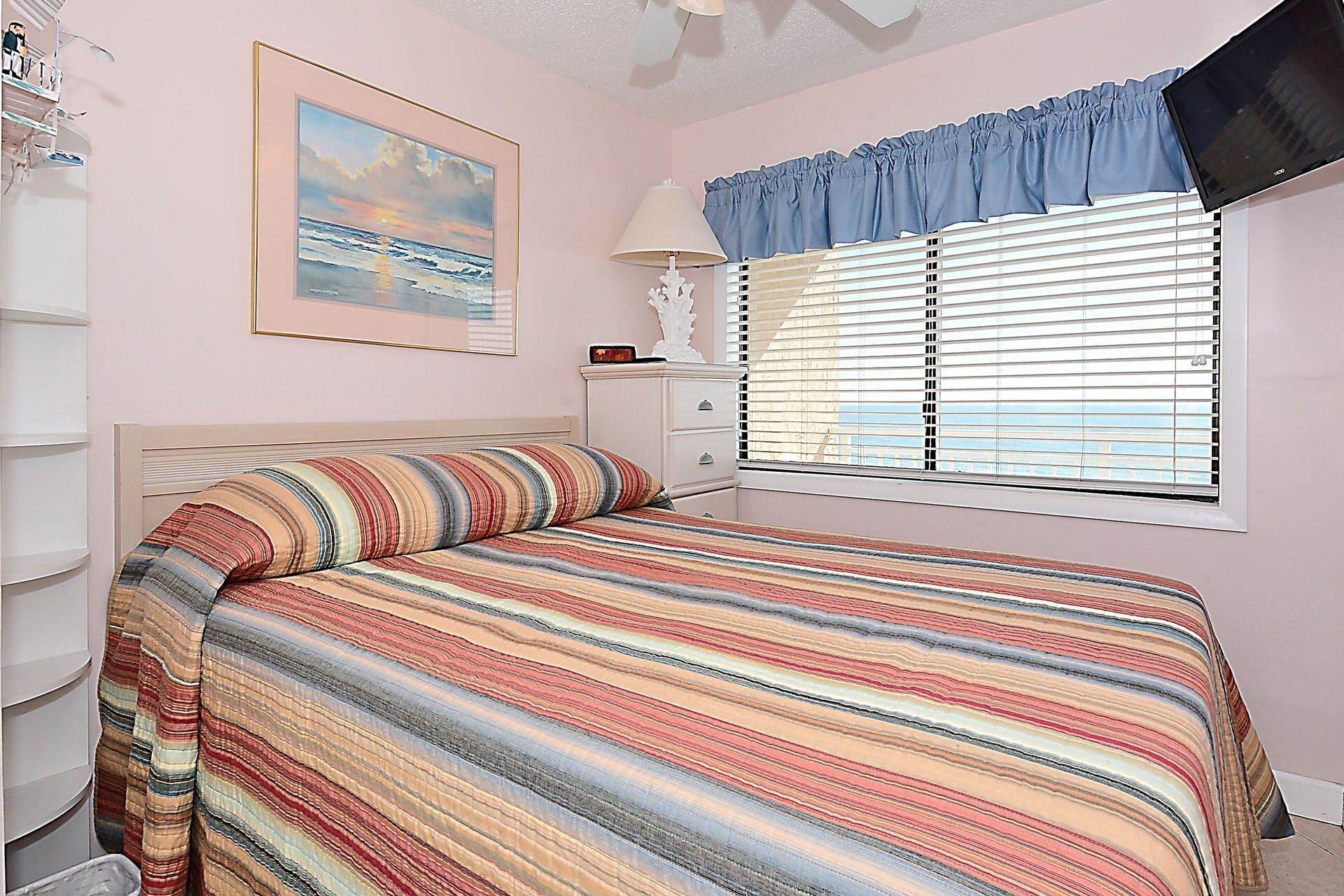 A bedroom with a bed, a window with blinds, a bedside table, and a wall-mounted TV.
