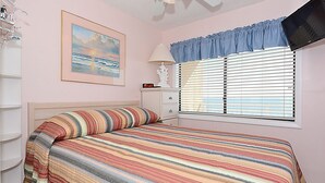 A bedroom with a bed, a window with blinds, a bedside table, and a wall-mounted TV.