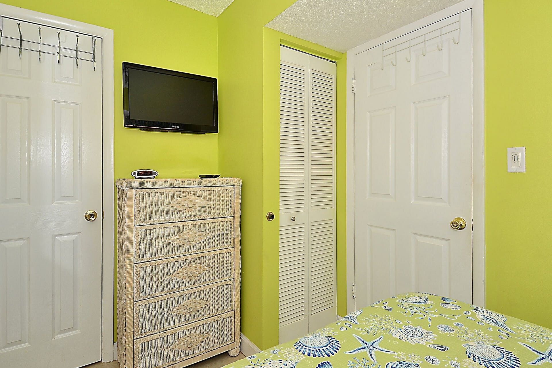 A room with two white doors, a wicker chest of drawers, a bed with a floral bedspread, and a flat-screen TV mounted on the wall.