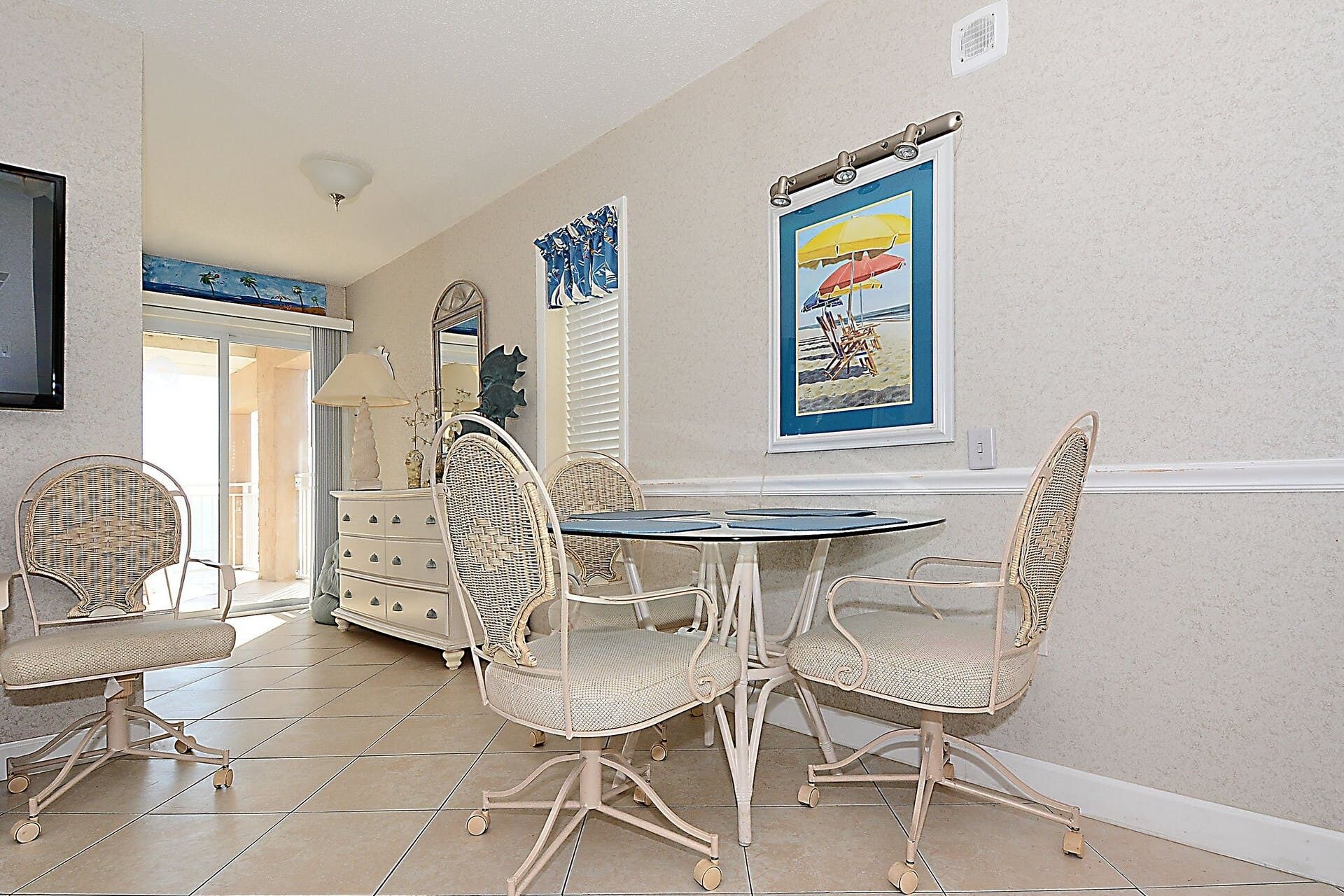 A dining area with wicker chairs and a round table, a white dresser, and a framed beach poster on the wall.