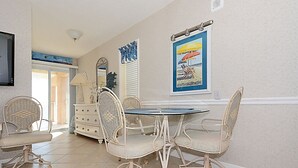 Un coin repas avec des chaises en osier et une table ronde, une commode blanche et une affiche représentant une plage, encadrée, accrochée au mur.