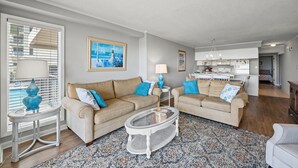 Ein Wohnzimmer mit zwei beigefarbenen Sofas, einem Couchtisch und einem gemusterten Teppich. An der Wand hängt ein Gemälde und ein Fenster mit Jalousien.