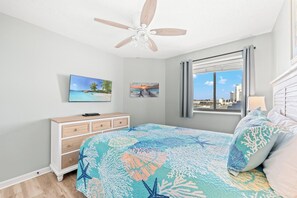 A bedroom with a bed, a ceiling fan, a dresser, a TV, and a window with a city view.