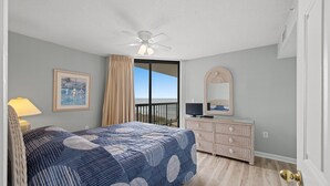 Ein Schlafzimmer mit einem Bett, einer Kommode, einem Deckenventilator, einem Fenster mit Vorhängen und einem gerahmten Bild an der Wand.