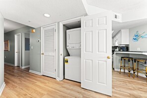 A laundry room with a washer and dryer, a kitchen with a bar stool, and a hallway with a door leading to another room. - 4-bedroom, 3-bathroom, oceanfront unit, sleeps 10 | Ocean Bay Club - 401 (North Myrtle Beach)