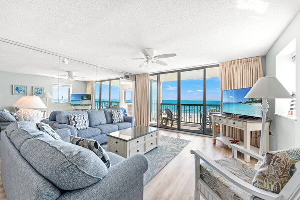 A modern living room with a sofa, coffee table, and TV, overlooking a balcony with a view of the ocean.