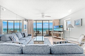 A spacious living room with a large sofa, a coffee table, and a TV. There is a balcony with a view of the ocean.