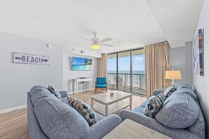 A living room with a sofa, a television, a ceiling fan, and a view of the beach through large windows. - 4-bedroom, 3-bathroom oceanfront condo with spacious private balcony - | Ocean Bay Club - 402 (North Myrtle Beach)