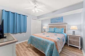 A bedroom with a bed, two bedside tables with lamps, a ceiling fan, a TV on a stand, and a door. - 4-bedroom, 3-bathroom oceanfront condo with spacious private balcony - | Ocean Bay Club - 402 (North Myrtle Beach)
