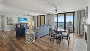 A spacious living room with a sofa, chairs, and a small table. There are two ceiling fans, a television, and a view of the ocean through large windows. - Oceanfront Penthouse, 4-bedrooms, 4-baths, balcony access from 2 bedrooms | Ocean Bay Club - 1702 PH (North Myrtle Beach)