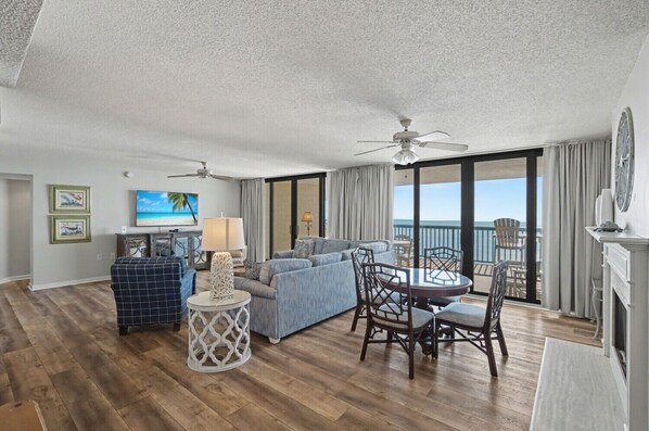 A spacious living room with a sofa, chairs, and a small table. There are two ceiling fans, a television, and a view of the ocean through large windows. - Oceanfront Penthouse, 4-bedrooms, 4-baths, balcony access from 2 bedrooms | Ocean Bay Club - 1702 PH (North Myrtle Beach)