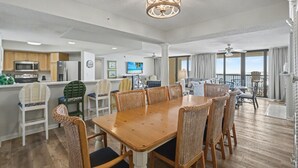 A spacious living area with a wooden dining table, wicker chairs, a kitchen with modern appliances, and a balcony with a view. - Oceanfront Penthouse, 4-bedrooms, 4-baths, balcony access from 2 bedrooms | Ocean Bay Club - 1702 PH (North Myrtle Beach)