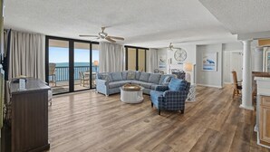 A spacious living room with a large sectional sofa, a coffee table, and a blue plaid armchair. There is a balcony with a view of the ocean. - Oceanfront Penthouse, 4-bedrooms, 4-baths, balcony access from 2 bedrooms | Ocean Bay Club - 1702 PH (North Myrtle Beach)