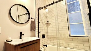 Une salle de bain avec un mur recouvert de carreaux blancs, un miroir rond, un meuble-lavabo en bois, un robinet noir, une cabine de douche en verre et une fenêtre.