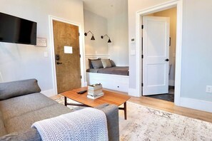 A modern living room with a sofa, a coffee table with books and a remote, a TV mounted on the wall, and a bed with a headboard in the background. - The Duke Suite at Prince Street Inn (Alexandria)