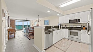A modern kitchen with white cabinets, a microwave, an oven, and a dishwasher. There is a dining area with a table and chairs, and a view of the ocean through large windows. - 3-bedroom oceanfront condo with lots of updates | Crescent Keyes - 705 (North Myrtle Beach)