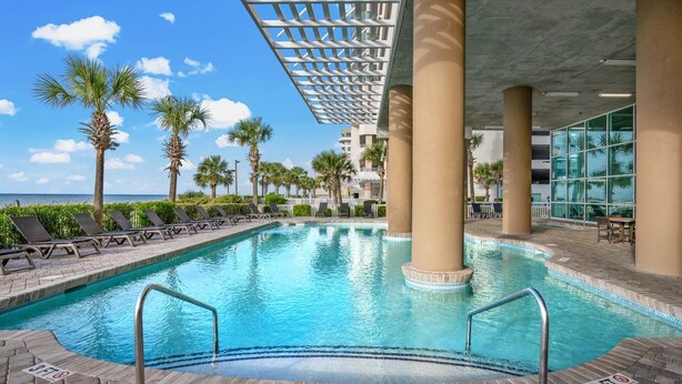 An indoor swimming pool with clear blue water, surrounded by lounge chairs and palm trees, under a covered area with a view of the ocean.