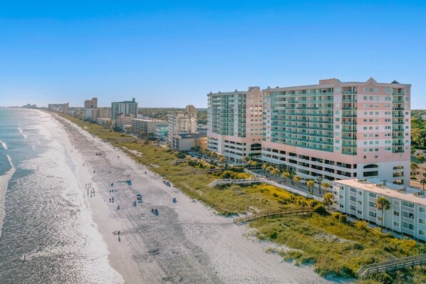 Un hotel frente a la playa con varios pisos, una playa en primer plano y un cielo despejado.