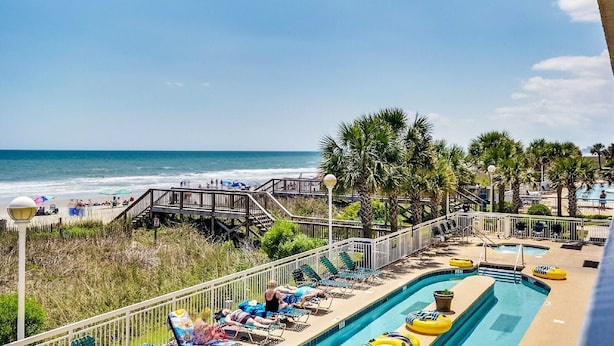 A pool area with lounge chairs, a wooden deck, and a view of the ocean. - Stylish 4-bedroom condo with jetted tub and two king bedrooms | Crescent Shores - N 1101 (North Myrtle Beach)