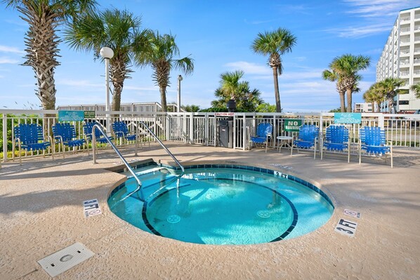 A hot tub surrounded by a pool, with blue chairs and palm trees in the background. - Stylish 4-bedroom condo with jetted tub and two king bedrooms | Crescent Shores - N 1101 (North Myrtle Beach)