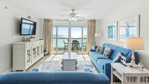 A living room with a blue sofa, a TV, a ceiling fan, and a view of the ocean through large windows.