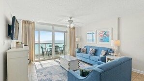 A living room with a blue sofa, a coffee table, and a TV. There is a balcony with a view of the sea.