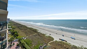 Vista da praia a partir de uma varanda, com céu claro, praia de areia e algumas pessoas caminhando.