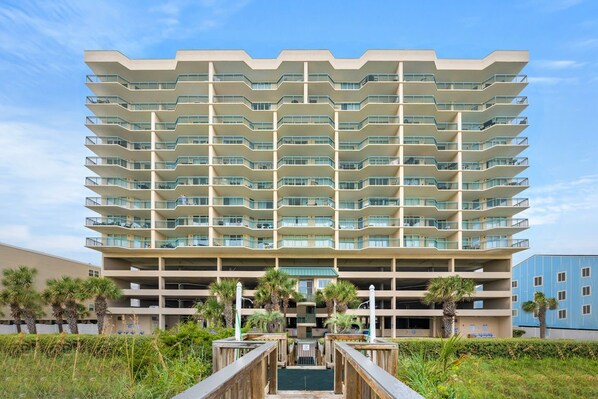 A multi-story hotel with a modern design, featuring balconies on each floor, a central courtyard, and a parking garage in the background.