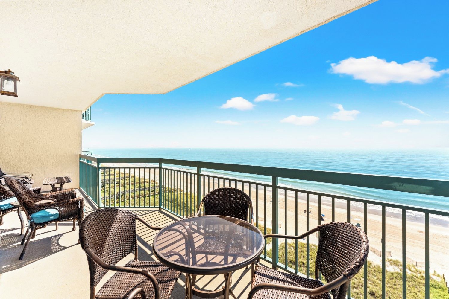 A balcony with wicker furniture and a glass table overlooking a beach.