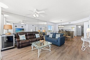 A spacious living area with a brown leather sofa, a blue sofa, and a glass coffee table. There is a dining area with a chandelier and a kitchen in the background. - Comfortable 4-bedroom, 3-bathroom oceanfront condo, Crescent Beach | North Shore Villas - 907 (North Myrtle Beach)