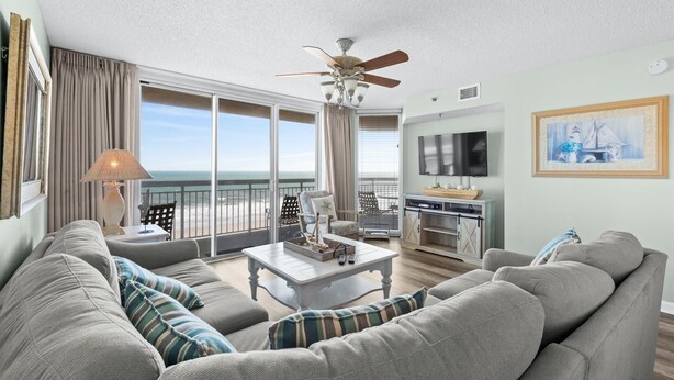 Ein geräumiges Wohnzimmer mit einer großen Couch, einem Couchtisch und einem Fernseher. Ein Balkon bietet Blick aufs Meer.