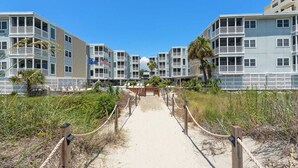 Een hotel aan het strand met meerlaagsgebouwen, een zandpad dat naar het strand leidt en een houten omheining.