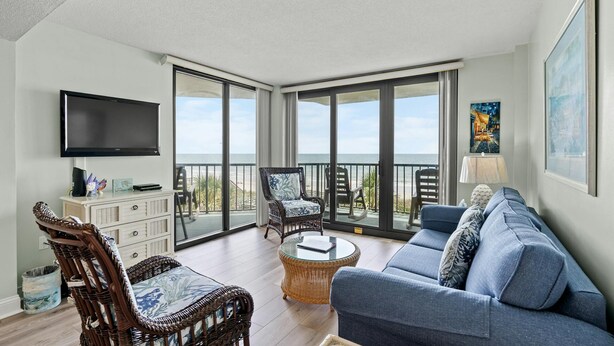 A spacious living room with a large TV, wicker furniture, a sofa, and a balcony with a view of the beach. - Classic beachfront condo with two beds and two baths, private balcony | Crescent Sands - 3-B (North Myrtle Beach)