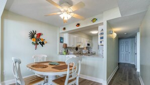 A dining area with a round wooden table and white chairs, a ceiling fan, and a kitchen in the background. - Classic beachfront condo with two beds and two baths, private balcony | Crescent Sands - 3-B (North Myrtle Beach)