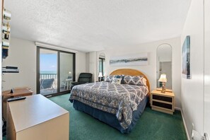A hotel room with a bed, a desk, a chair, a nightstand, a lamp, a painting, and a view of the ocean. - 3-bedroom oceanfront condo with private balcony | Crescent Sands - 5-G (North Myrtle Beach)