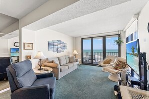 A living room with a sofa, armchairs, a TV, and a large window with a view of the beach. - 3-bedroom oceanfront condo with private balcony | Crescent Sands - 5-G (North Myrtle Beach)
