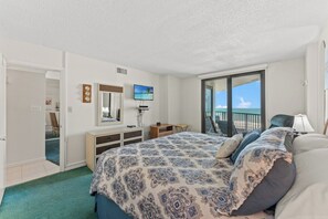 A bedroom with a bed, a dresser, a TV, a balcony with a view of the ocean, and a door leading to another room.