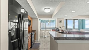Une cuisine moderne équipée d’un réfrigérateur, d’une cuisinière et d’un évier, un coin repas avec une chaise, et une vue sur l’océan grâce à de grandes fenêtres.