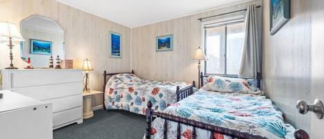 A bedroom with two beds, a dresser, a window with curtains, and two wall-mounted paintings.