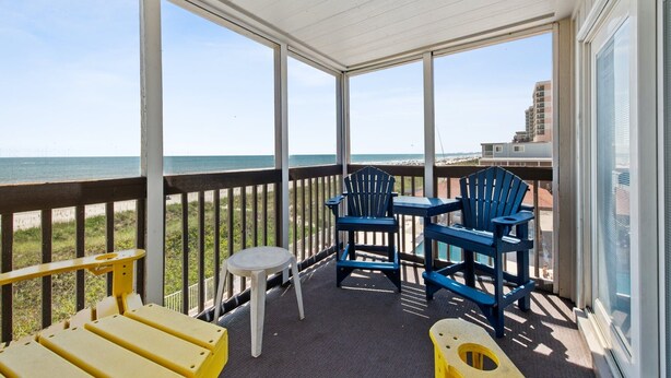 A covered balcony with a view of the beach and ocean, featuring blue and yellow Adirondack chairs and a small white table. - Two bedroom, two-bathroom oceanfront condo | Crescent Beach Villas - 201 (North Myrtle Beach)