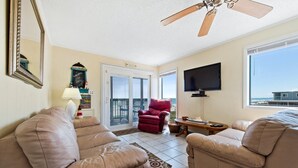 A living room with two beige sofas, a red chair, a television, and a ceiling fan. - Two bedroom, two-bathroom oceanfront condo | Crescent Beach Villas - 201 (North Myrtle Beach)