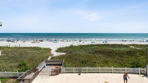 Une plage avec une promenade en bois menant à une zone sablonneuse, une clôture blanche en piquets et un panneau indiquant qu’il n’y a pas de sauveteur en service.